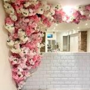 May include: A white brick wall with a pink and white floral arrangement in the corner. The flowers are cascading down the wall and are made of silk.