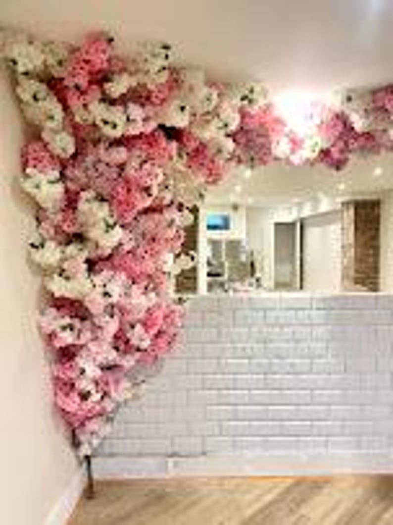 May include: A white brick wall with a pink and white floral arrangement in the corner. The flowers are cascading down the wall and are made of silk.