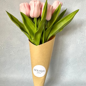 Op de afbeelding: Een boeket roze tulpen met groene bladeren, verpakt in een bruine papieren kegel. De bloemen hebben een zacht, delicaat uiterlijk. Een witte ronde sticker met de tekst "SDesign Floral" is op het papier bevestigd.