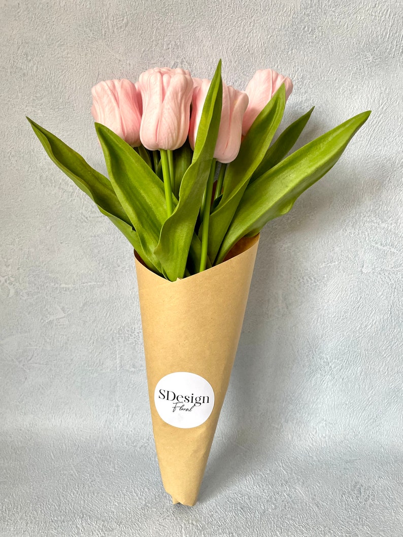 Op de afbeelding: Een boeket roze tulpen met groene bladeren, verpakt in een bruine papieren kegel. De bloemen hebben een zacht, delicaat uiterlijk. Een witte ronde sticker met de tekst "SDesign Floral" is op het papier bevestigd.