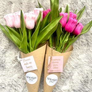 Op de afbeelding: Twee boeketten tulpen, &eacute;&eacute;n lichtroze en &eacute;&eacute;n felroze, verpakt in bruine papieren kegels. Elk boeket heeft een label met de tekst "Flowers make me happy" en "happiness blooms from within". De boeketten zijn van SDesign Floral.