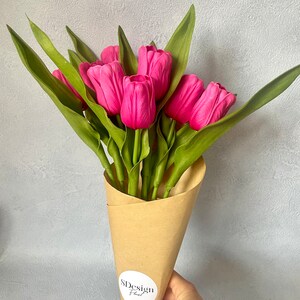 Op de afbeelding: Een boeket van felroze tulpen met groene bladeren, verpakt in een bruine papieren kegel. Het boeket wordt door een hand vastgehouden. Een witte sticker met de tekst "SDesign Floral" staat op de papieren kegel.