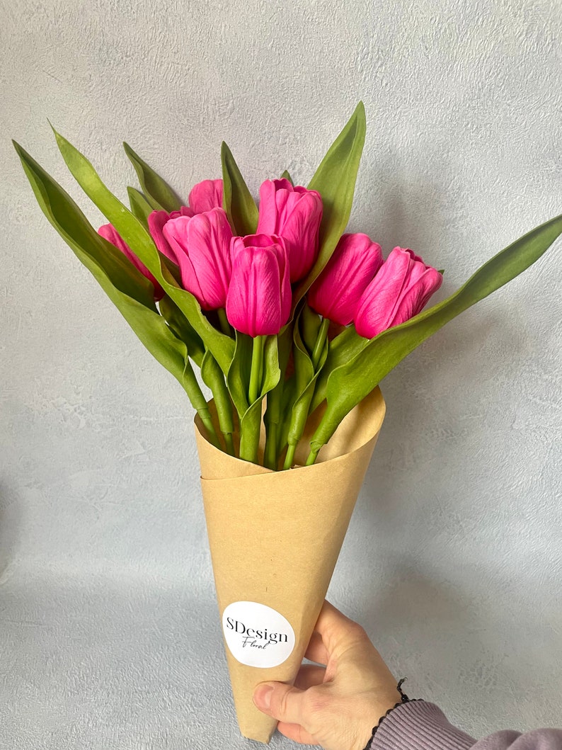 Op de afbeelding: Een boeket van felroze tulpen met groene bladeren, verpakt in een bruine papieren kegel. Het boeket wordt door een hand vastgehouden. Een witte sticker met de tekst "SDesign Floral" staat op de papieren kegel.