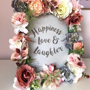 May include: A round, gray picture frame with a white background and the text "Happiness Love & Laughter" in silver glitter. The frame is decorated with a wreath of pink, white, and gray artificial flowers.