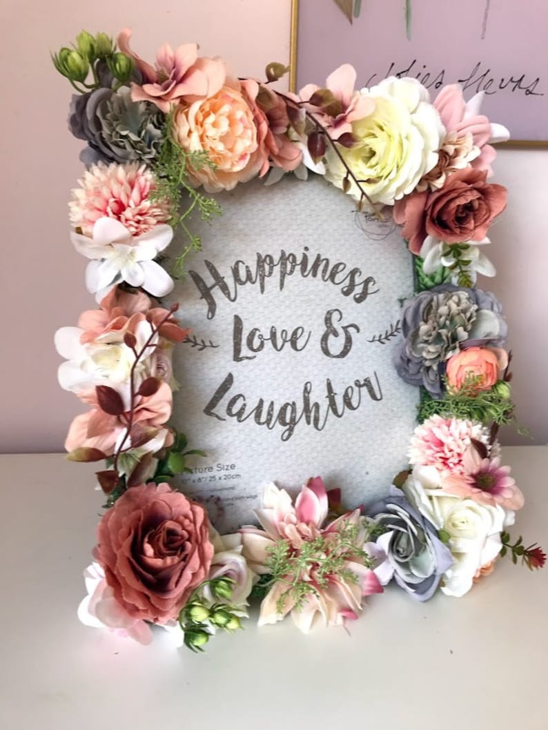 May include: A round, gray picture frame with a white background and the text "Happiness Love & Laughter" in silver glitter. The frame is decorated with a wreath of pink, white, and gray artificial flowers.