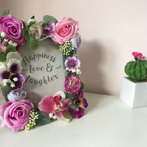 May include: A floral picture frame with a pink, purple, and white flower arrangement. The frame has a sign that reads "Happiness Love & Laughter".