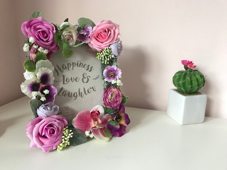 May include: A floral picture frame with a pink, purple, and white flower arrangement. The frame has a sign that reads "Happiness Love & Laughter".