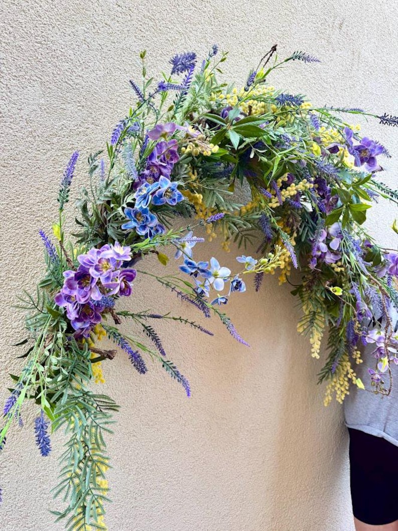 May include: A colourful floral archway arrangement with purple, blue, and yellow flowers and green leaves. The flowers include lavender and orchids. Ideal for weddings or home decoration. The arrangement is in an arch shape.