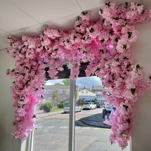 May include: A window display decorated with a large pink floral arrangement of artificial cherry blossoms. The flowers are cascading down from the ceiling, creating a beautiful and whimsical effect.
