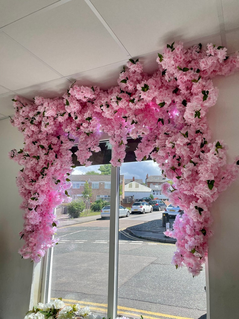 May include: A window display decorated with a large pink floral arrangement of artificial cherry blossoms. The flowers are cascading down from the ceiling, creating a beautiful and whimsical effect.