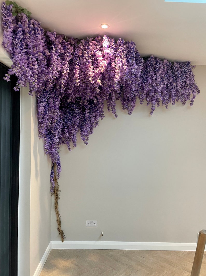 May include: A corner of a room decorated with a long strand of artificial purple wisteria flowers hanging from the ceiling.