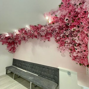 May include: A gray tufted bench with silver legs sits in front of a pink wall decorated with a large floral arrangement of pink blossoms.
