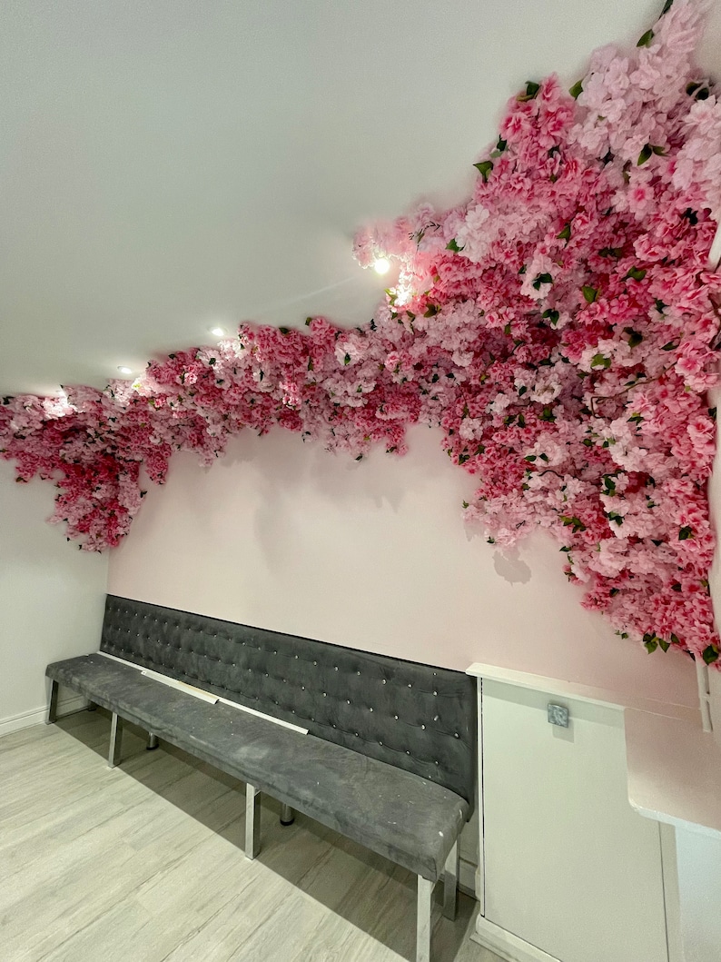 May include: A gray tufted bench with silver legs sits in front of a pink wall decorated with a large floral arrangement of pink blossoms.