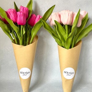 Op de afbeelding: Twee boeketten tulpen in bruine papieren kegels. E&eacute;n boeket heeft felroze tulpen, de andere zachtroze. Beide hebben groene bladeren en witte ronde stickers met "SDesign Floral" erop.