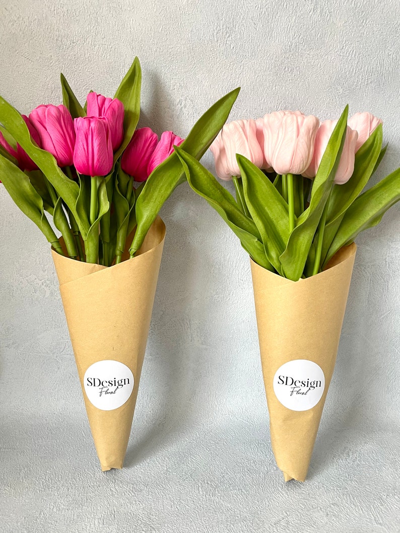 Op de afbeelding: Twee boeketten tulpen in bruine papieren kegels. E&eacute;n boeket heeft felroze tulpen, de andere zachtroze. Beide hebben groene bladeren en witte ronde stickers met "SDesign Floral" erop.