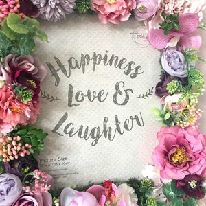 May include: A white picture frame with a floral border made of pink, purple, and green artificial flowers. The frame has the text "Happiness Love & Laughter" and "Picture Size 10" x 8"/25 x 20cm" printed on it.