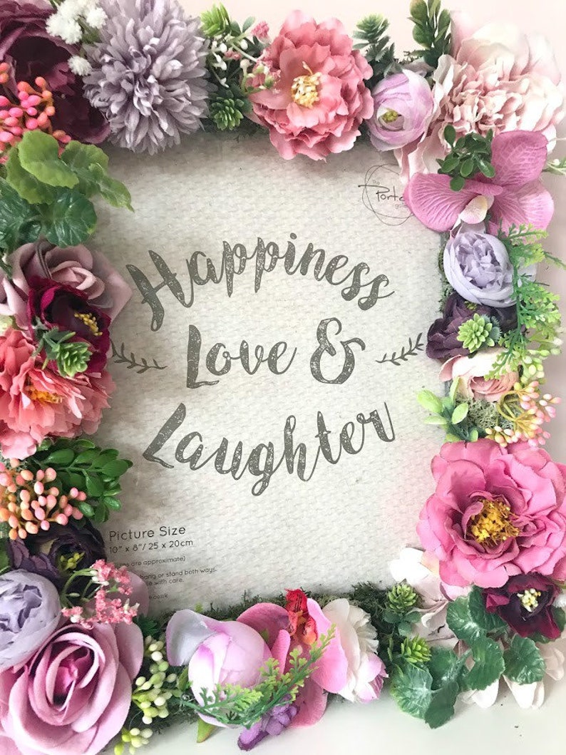 May include: A white picture frame with a floral border made of pink, purple, and green artificial flowers. The frame has the text "Happiness Love & Laughter" and "Picture Size 10" x 8"/25 x 20cm" printed on it.
