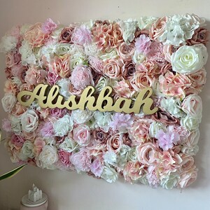 May include: A pink and white flower wall with the name "Alishbah" written in gold letters.