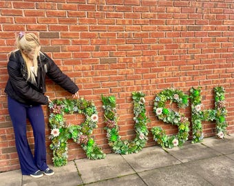 Puede incluir: Una mujer se para frente a una pared de ladrillo con la palabra "RUSH" escrita en letras grandes hechas de plantas verdes.