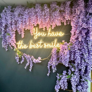 May include: A neon sign with the text "You have the best smile" hanging on a wall decorated with purple wisteria flowers. A brown leather bench sits in front of the wall.