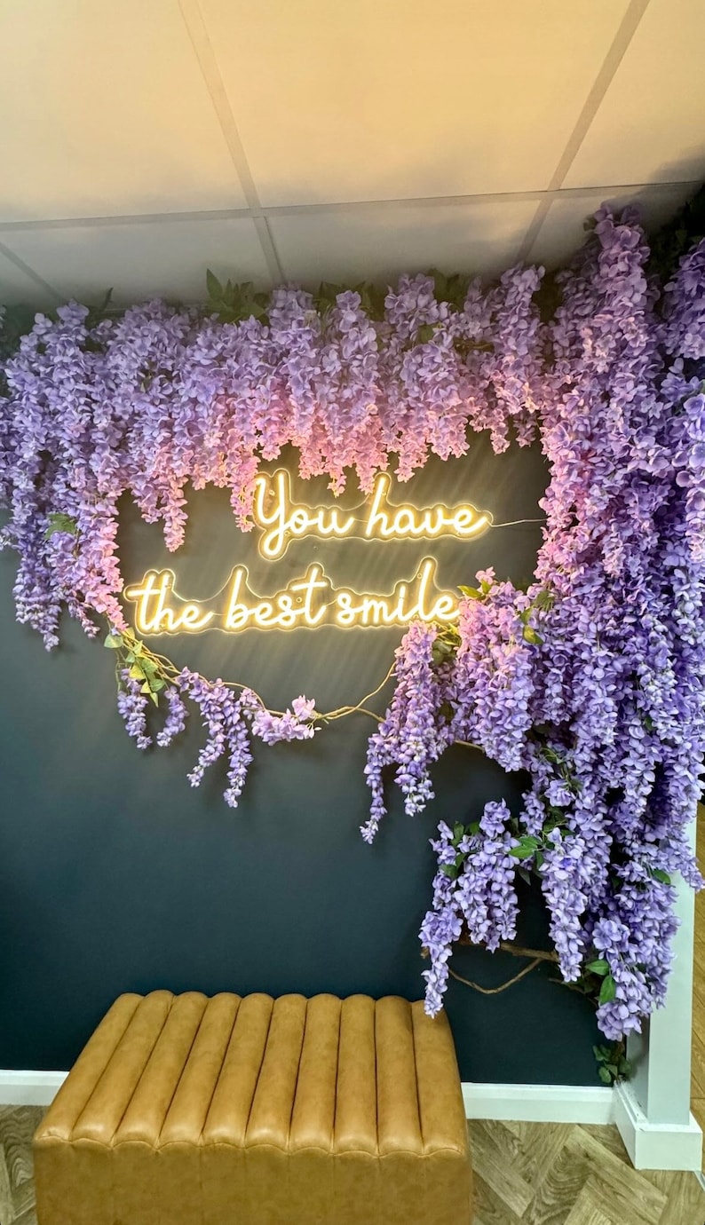 May include: A neon sign with the text "You have the best smile" hanging on a wall decorated with purple wisteria flowers. A brown leather bench sits in front of the wall.