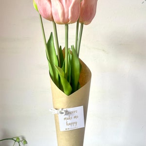 May include: A bouquet of pink tulips wrapped in brown paper with a white tag that says "Flowers make me happy". The bouquet is held by a hand with a white sticker on the wrist that says "SDesign Floral".