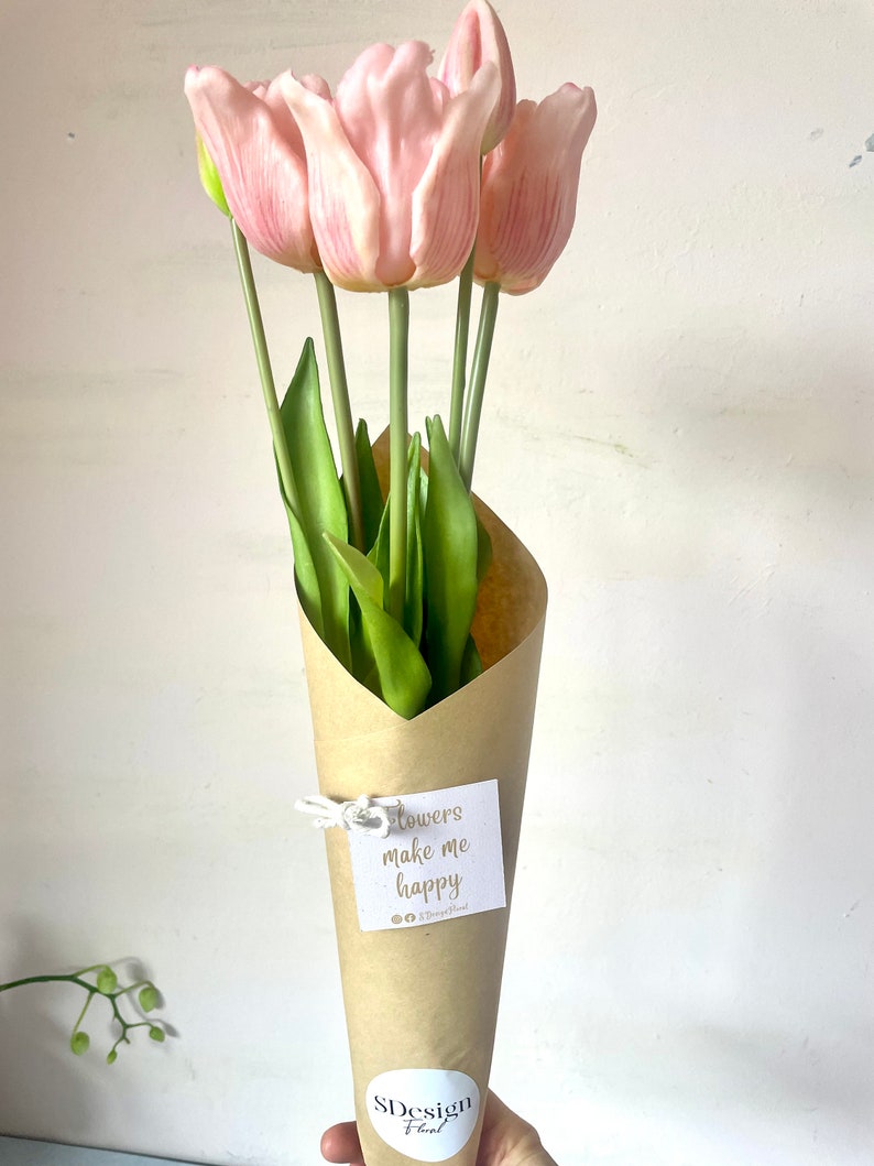 May include: A bouquet of pink tulips wrapped in brown paper with a white tag that says "Flowers make me happy". The bouquet is held by a hand with a white sticker on the wrist that says "SDesign Floral".