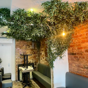 Peut inclure: Int&eacute;rieur de restaurant avec un mur en brique, une banquette grise et une petite table avec deux chaises. Le restaurant est d&eacute;cor&eacute; de verdure artificielle suspendue au plafond.