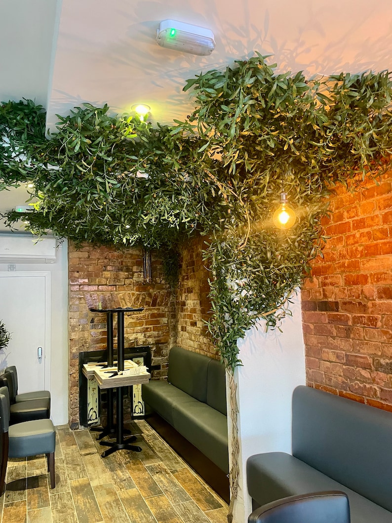 Peut inclure: Int&eacute;rieur de restaurant avec un mur en brique, une banquette grise et une petite table avec deux chaises. Le restaurant est d&eacute;cor&eacute; de verdure artificielle suspendue au plafond.