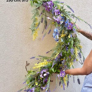 May include: A floral garland, approximately 5 feet (150 cm) long, featuring purple, yellow, and blue flowers with green foliage. The arrangement is held against a neutral-colored wall, showcasing its cascading design.