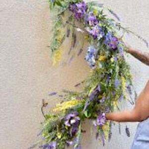 May include: A floral arrangement featuring purple, lavender, and yellow flowers with green foliage. The arrangement is being held against a light-colored wall. The flowers are a mix of different shapes and sizes, creating a natural, organic look.
