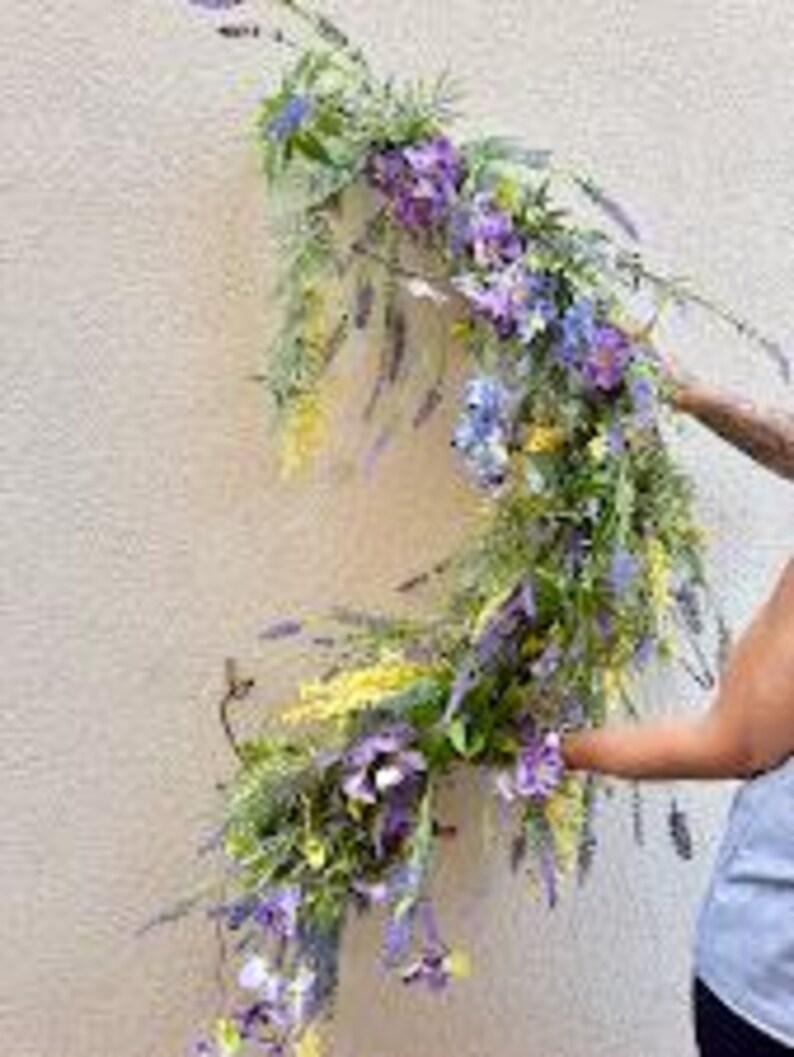 May include: A floral arrangement featuring purple, lavender, and yellow flowers with green foliage. The arrangement is being held against a light-colored wall. The flowers are a mix of different shapes and sizes, creating a natural, organic look.