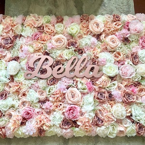 May include: A flower wall backdrop featuring a mix of pink, white and cream artificial flowers. The name "Bella" is written in cursive on a wooden sign in the centre of the wall.