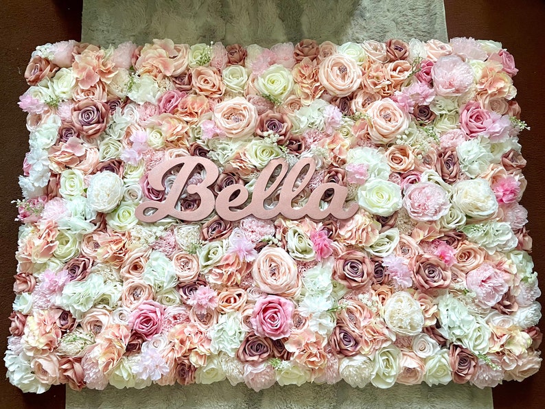 May include: A flower wall backdrop featuring a mix of pink, white and cream artificial flowers. The name "Bella" is written in cursive on a wooden sign in the centre of the wall.
