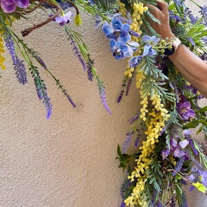 May include: A vibrant floral arrangement featuring purple, blue, and yellow flowers. The arrangement includes lavender, orchids, and other greenery. The flowers are arranged in a semi-circular shape, with a hand holding the arrangement.