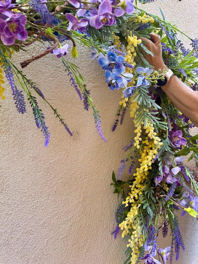 May include: A vibrant floral arrangement featuring purple, blue, and yellow flowers. The arrangement includes lavender, orchids, and other greenery. The flowers are arranged in a semi-circular shape, with a hand holding the arrangement.