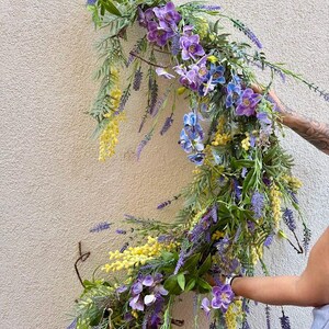 May include: A vibrant floral arrangement featuring a cascade of purple, blue, and yellow flowers. The arrangement includes lavender, orchids, and other greenery, creating a natural and colorful display. The flowers are arranged on a curved branch.