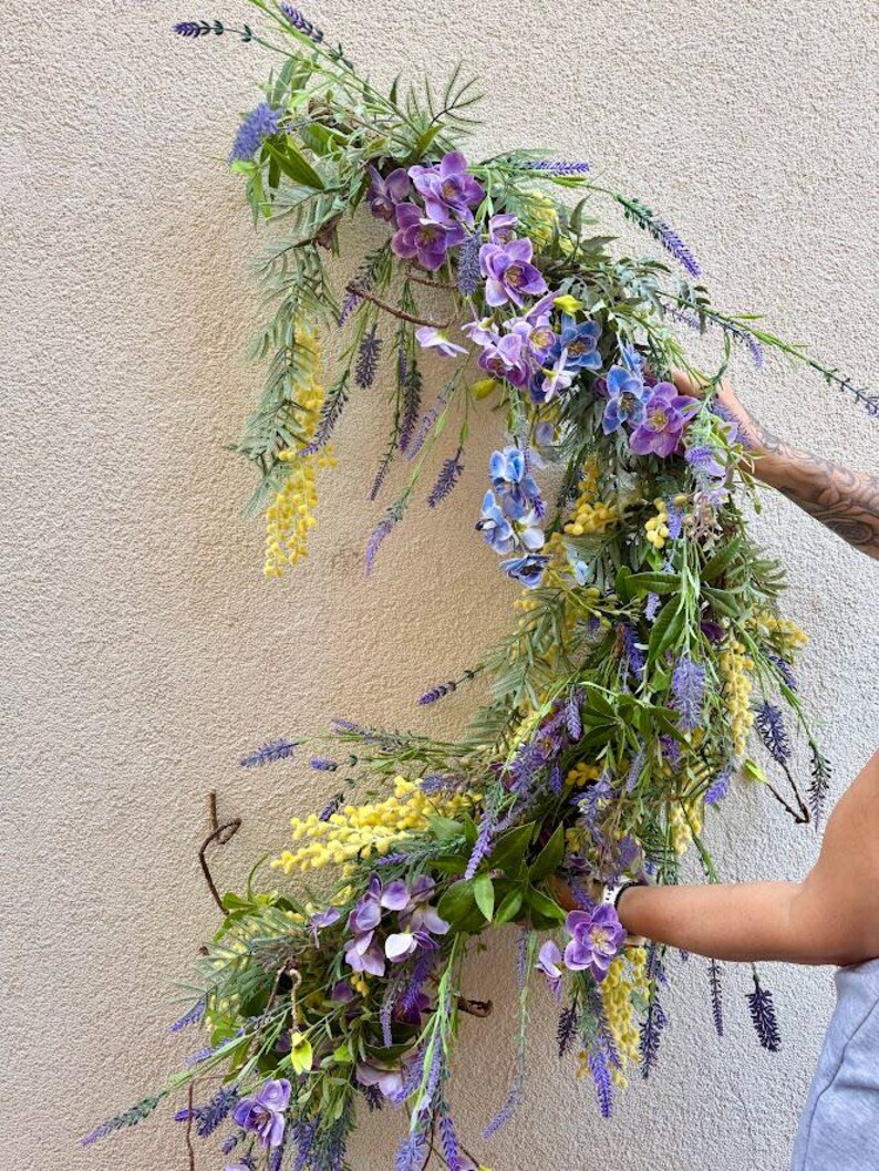 May include: A vibrant floral arrangement featuring a cascade of purple, blue, and yellow flowers. The arrangement includes lavender, orchids, and other greenery, creating a natural and colorful display. The flowers are arranged on a curved branch.