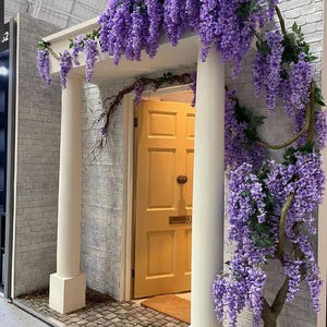 May include: A white brick building with a yellow door and a purple wisteria vine growing over the entrance. The vine is in full bloom and the flowers are cascading down the side of the building.