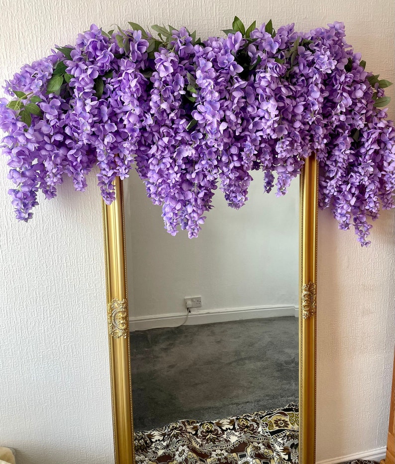 May include: A gold framed mirror with a purple wisteria garland draped over the top. The mirror is reflecting a carpeted floor and a patterned rug.