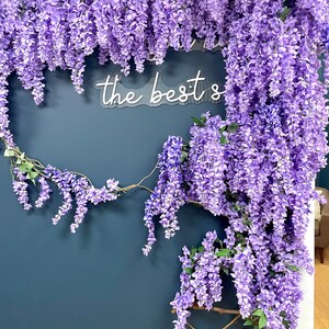 May include: A dark blue wall decorated with a garland of purple wisteria flowers. A white neon sign with the text "the best's" is mounted on the wall.