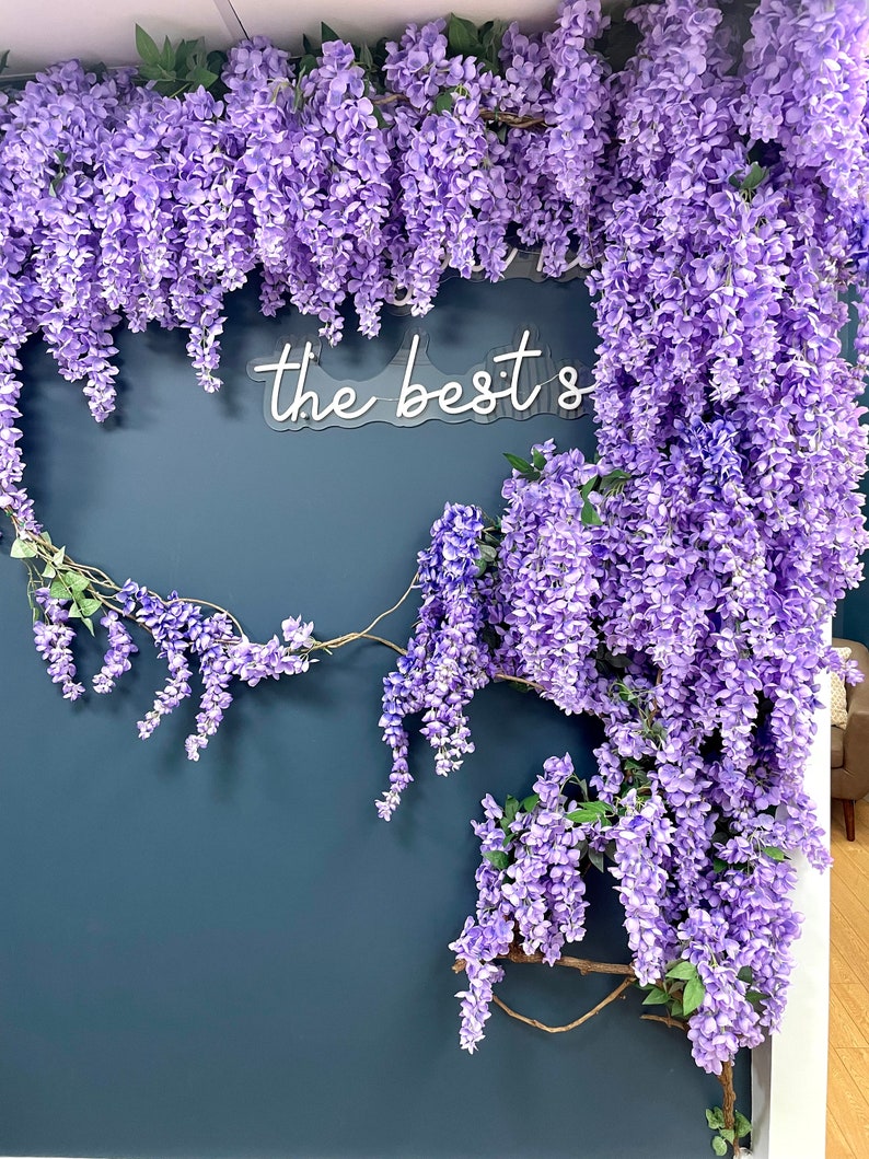 May include: A dark blue wall decorated with a garland of purple wisteria flowers. A white neon sign with the text "the best's" is mounted on the wall.