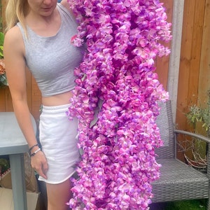May include: A woman is holding a large, long strand of artificial wisteria flowers. The flowers are a vibrant purple and pink color. The woman is wearing a grey tank top, white shorts, and white socks with black stripes and the words "new balance" written on them.