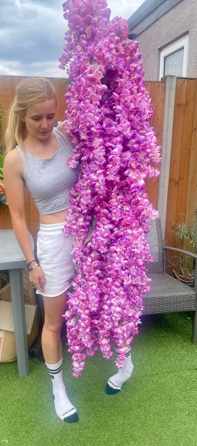 May include: A woman is holding a large, long strand of artificial wisteria flowers. The flowers are a vibrant purple and pink color. The woman is wearing a grey tank top, white shorts, and white socks with black stripes and the words "new balance" written on them.