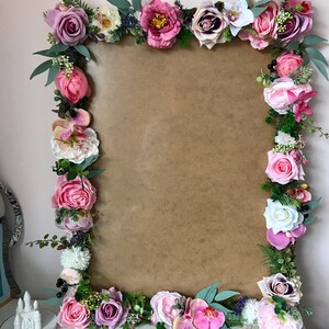 May include: A brown rectangular frame decorated with a floral garland of pink, purple, and white flowers. The garland includes roses, peonies, orchids, and greenery.