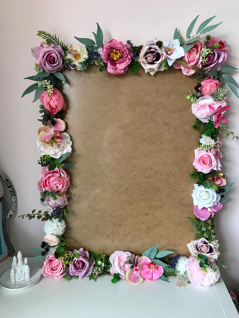 May include: A brown rectangular frame decorated with a floral garland of pink, purple, and white flowers. The garland includes roses, peonies, orchids, and greenery.