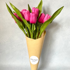 Op de afbeelding: Een boeket van felroze tulpen met groene bladeren, gepresenteerd in een bruine papieren kegel. De bloemen zijn dicht bij elkaar geclusterd, waardoor een volle, weelderige uitstraling ontstaat. Een witte sticker met de tekst "SDesign Floral" is op de papieren kegel bevestigd.