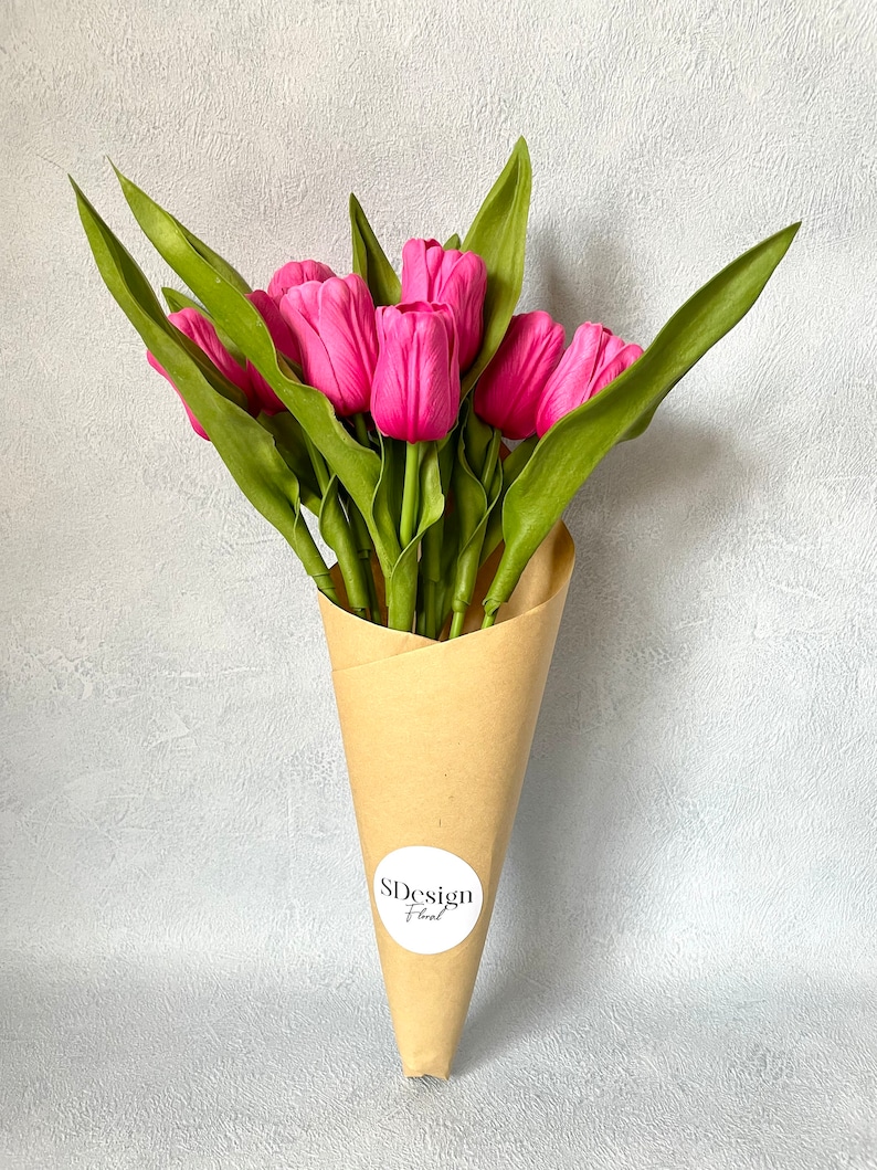 Op de afbeelding: Een boeket van felroze tulpen met groene bladeren, gepresenteerd in een bruine papieren kegel. De bloemen zijn dicht bij elkaar geclusterd, waardoor een volle, weelderige uitstraling ontstaat. Een witte sticker met de tekst "SDesign Floral" is op de papieren kegel bevestigd.