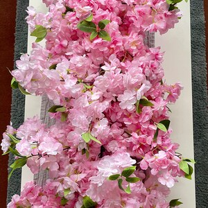 May include: A long garland of pink artificial cherry blossoms with green leaves. The garland is draped over a white surface.