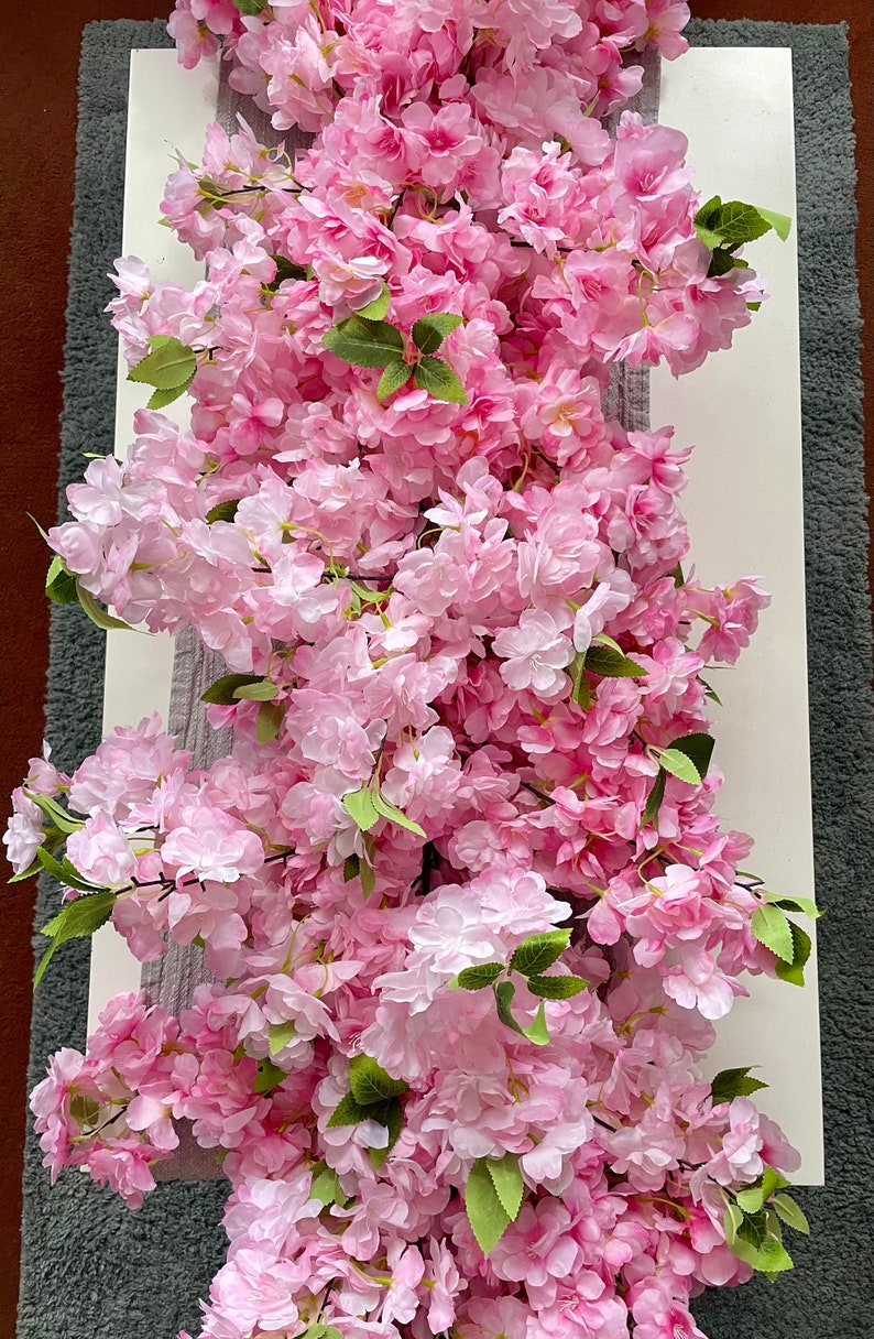 May include: A long garland of pink artificial cherry blossoms with green leaves. The garland is draped over a white surface.